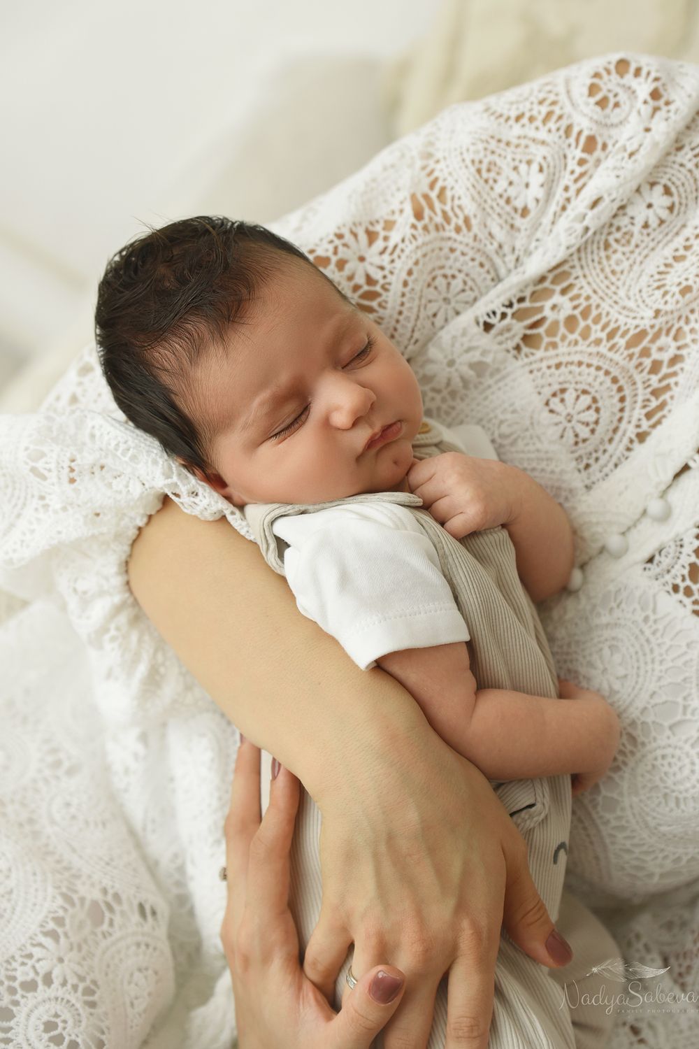 A sleeping newborn baby rests peacefully against white lace bedding while holding someone's hand.