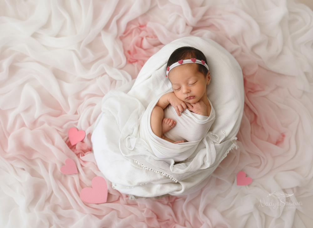 A sleeping newborn baby wrapped in white swaddle cloth lying on pink textured fabric with scattered paper hearts.