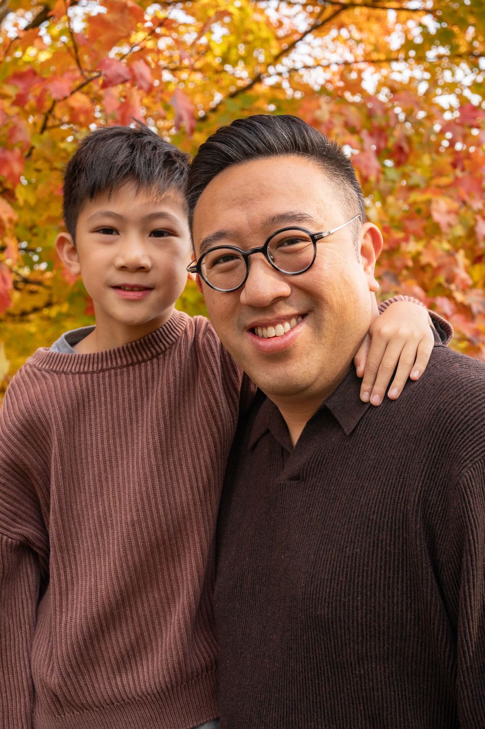 Autumn family portrait with warm brown sweaters against vibrant fall foliage background.
