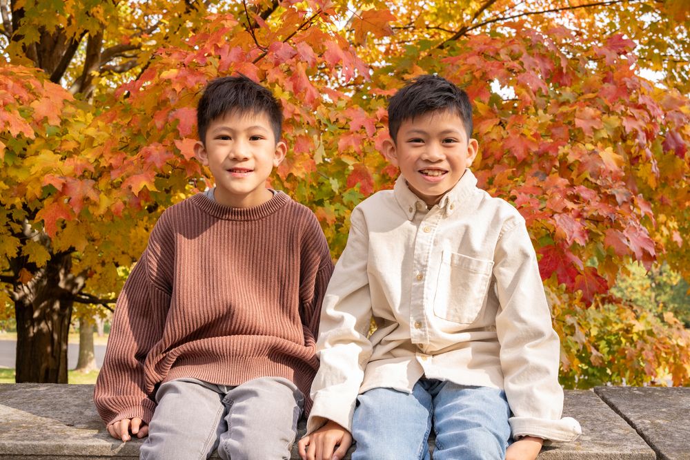 Two children sit together on a bench surrounded by vibrant autumn maple trees with red and orange leaves.