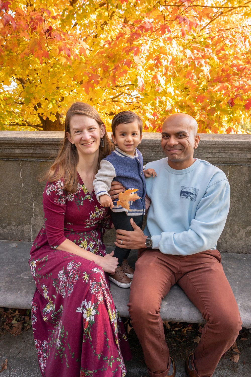 A family sits together on a stone wall with vibrant orange and yellow autumn foliage in the background.