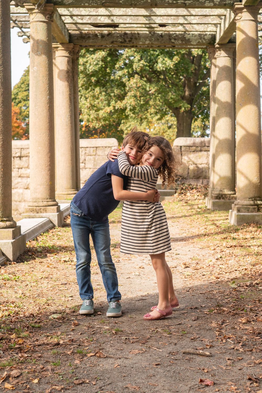 A playful moment under a stone pergola with autumn leaves scattered on the ground.