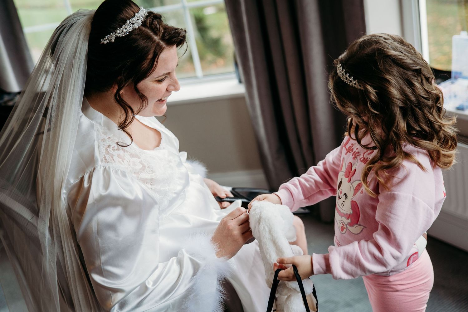A person in a wedding dress gets assistance with final preparations from someone wearing pink.