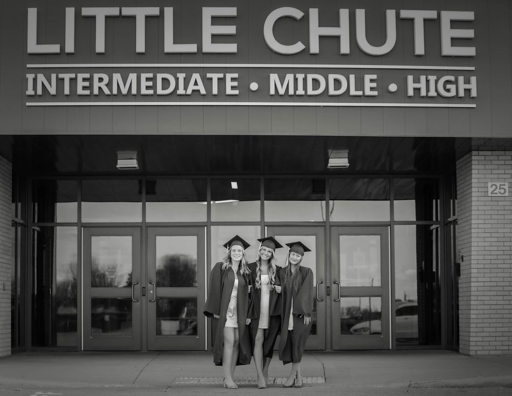 A black and white photo of graduates in caps and gowns standing in front of Little Chute Intermediate-Middle-High School entrance.
