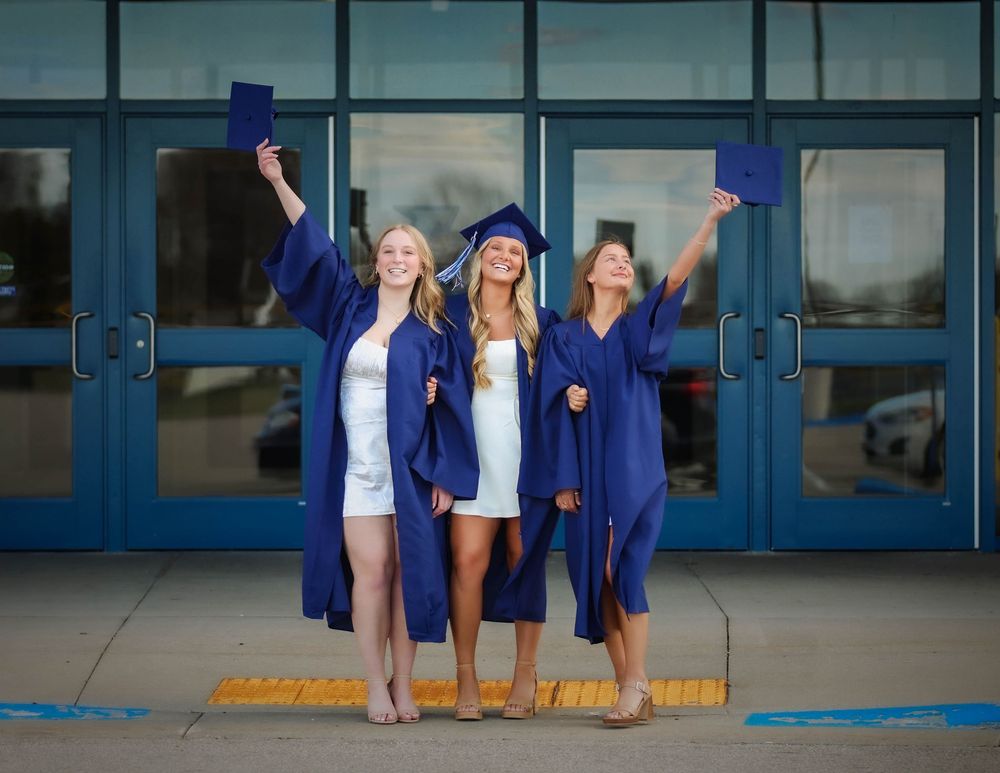 Three graduates in blue caps and gowns celebrate together outside glass doors while holding up their diplomas.