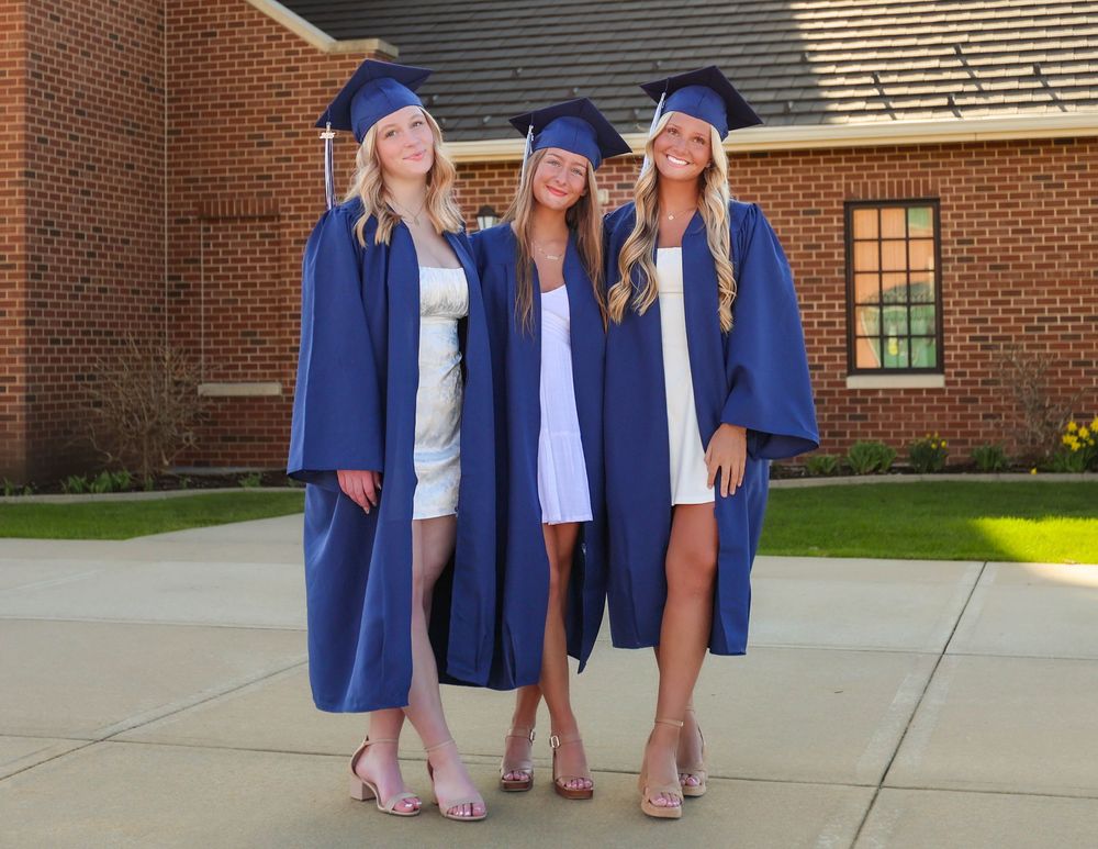 Three graduates in matching blue caps and gowns pose together on a sunny day outside a brick building.