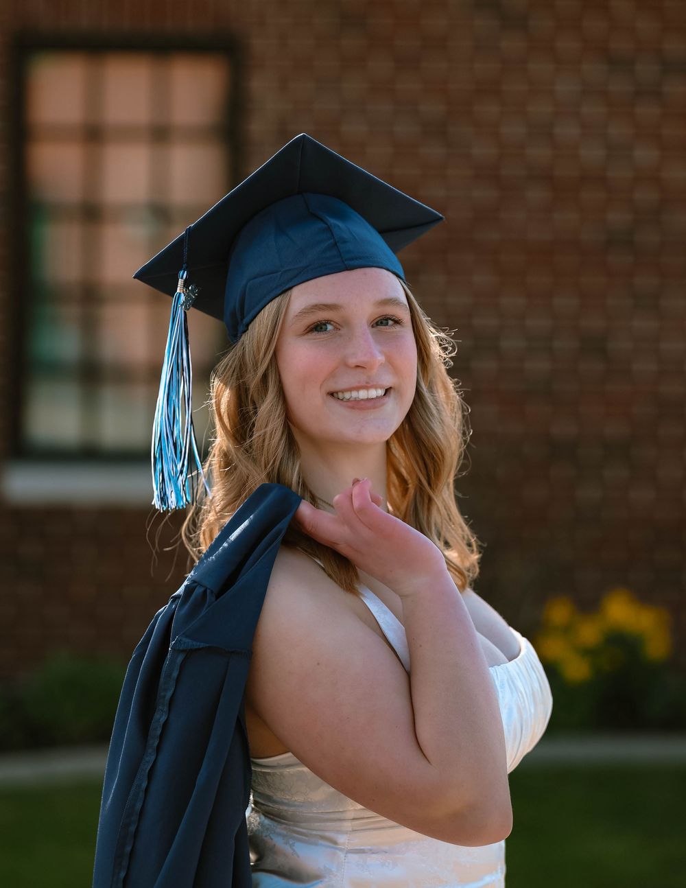 Graduate in cap and gown poses outdoors against brick building on graduation day.