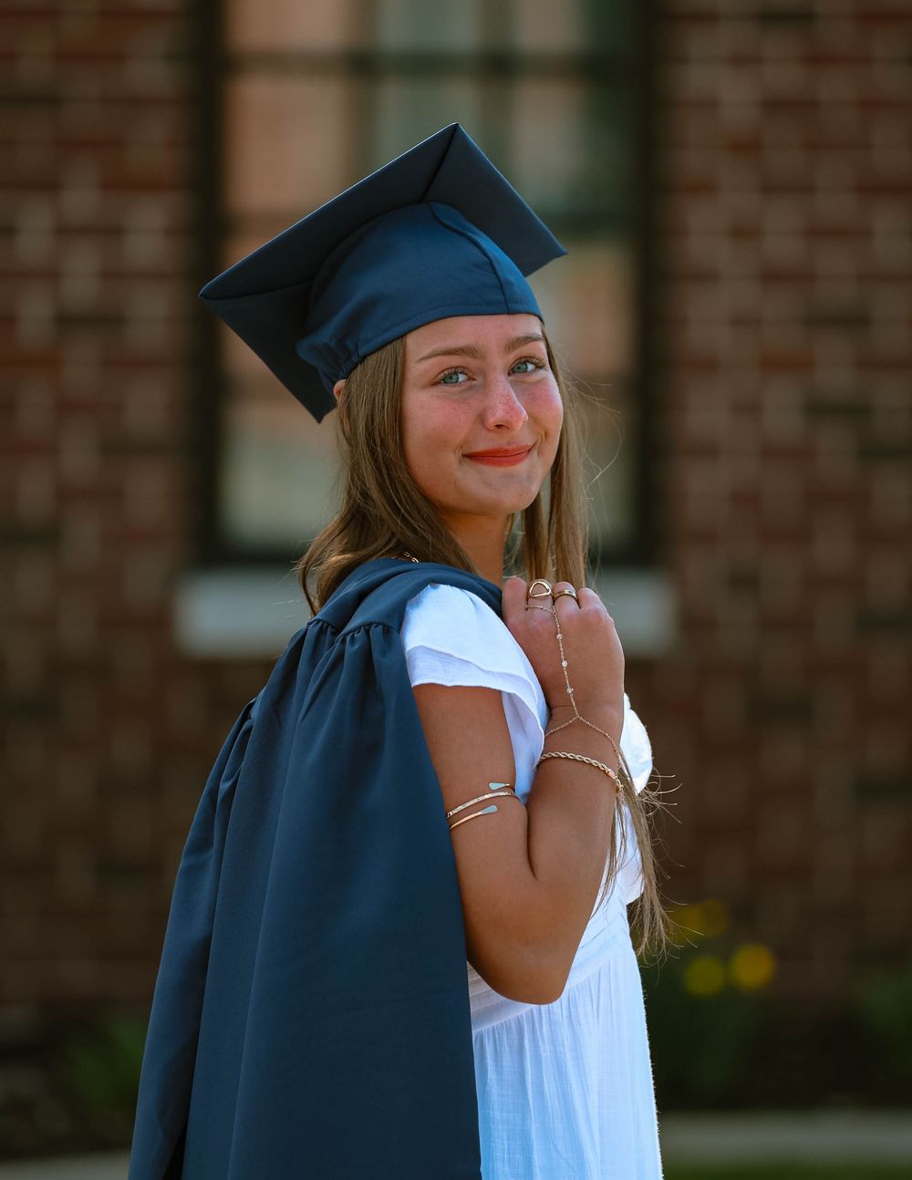 Graduate in navy cap and gown smiles over shoulder while posing in front of brick building during sunny day.