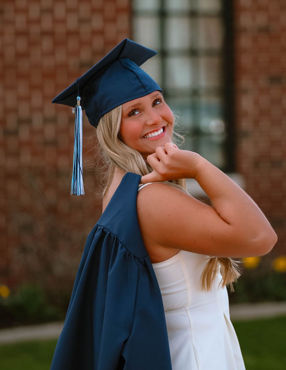 A graduate wearing a navy blue cap and gown smiles while posing in front of a brick building on graduation day.