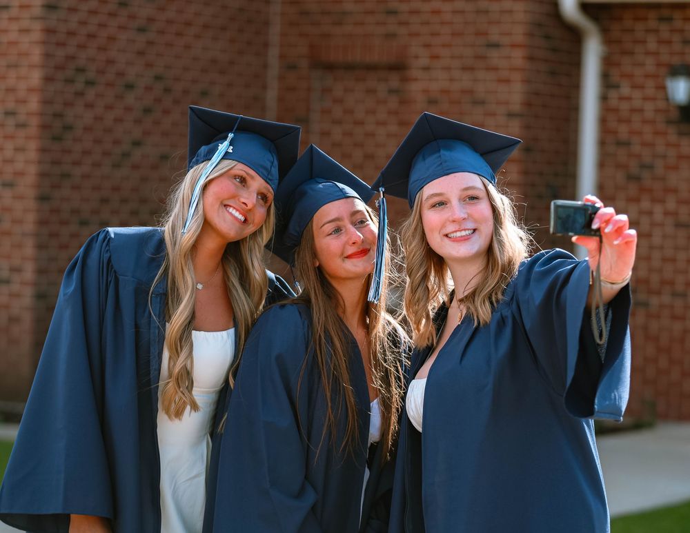 Three graduates in navy blue caps and gowns celebrate together on graduation day against a brick wall background.