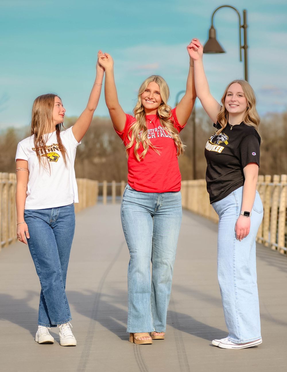 Three friends wearing collegiate shirts and jeans hold hands while celebrating together on an outdoor walkway.