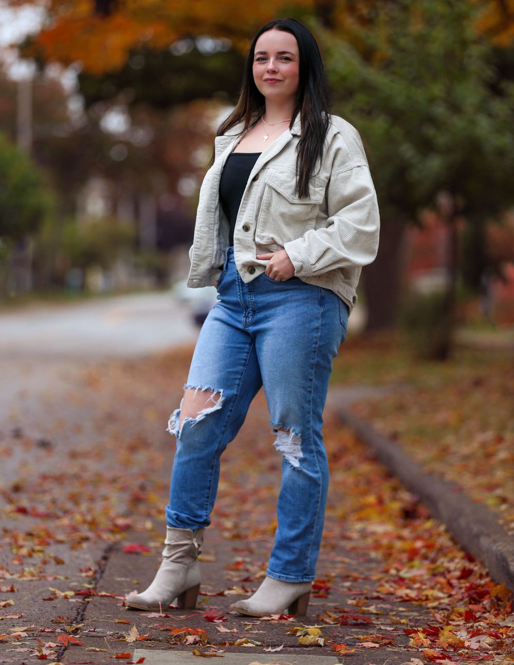 Fall fashion portrait of someone in ripped jeans, boots and white jacket standing among autumn leaves.