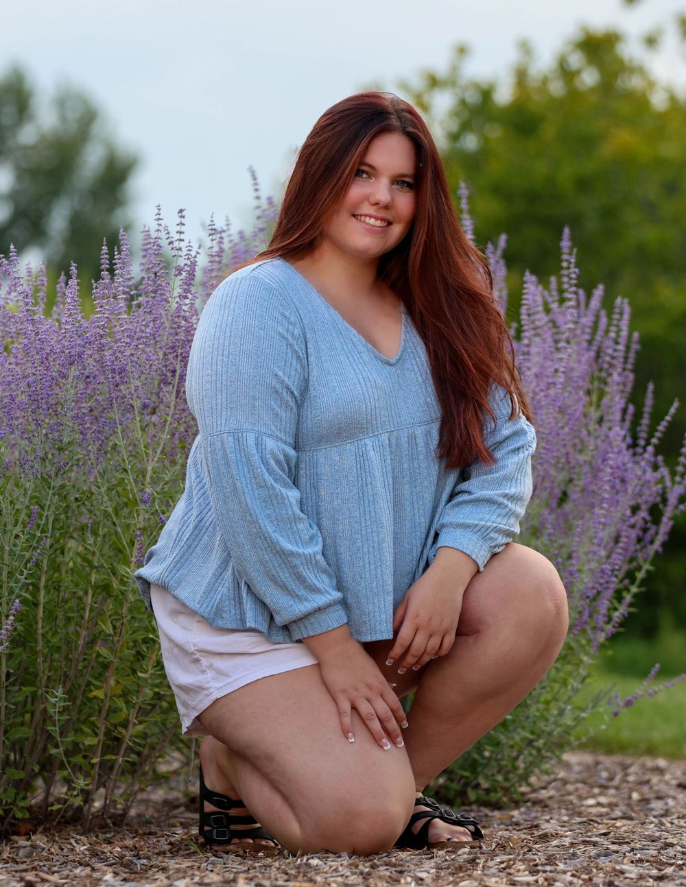 Woman in blue sweater kneels beside blooming purple lavender plants.