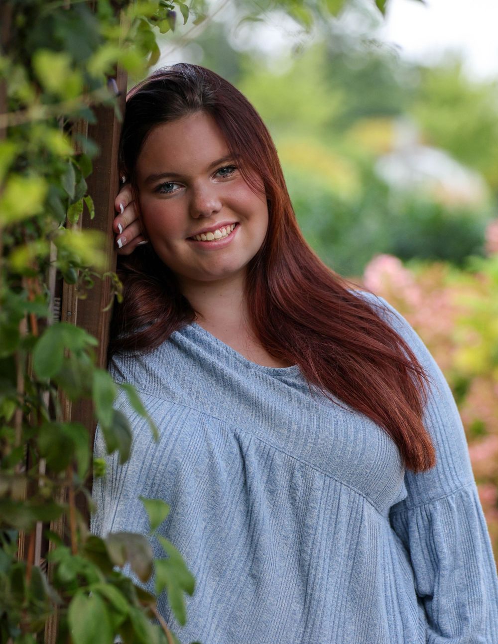 Woman in light blue top poses against garden greenery backdrop.