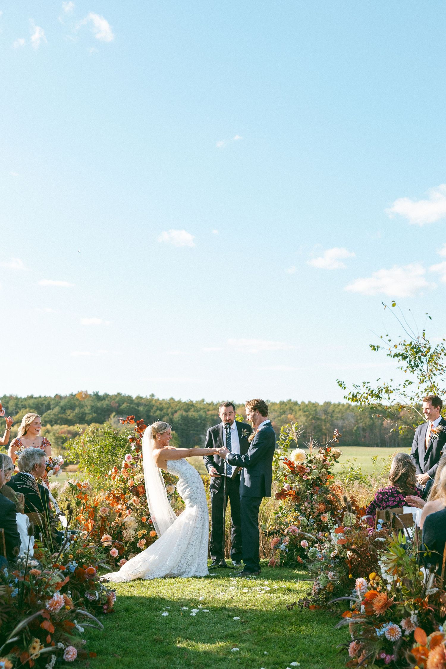 Outdoor wedding ceremony in a scenic field with fall foliage and floral arrangements.
