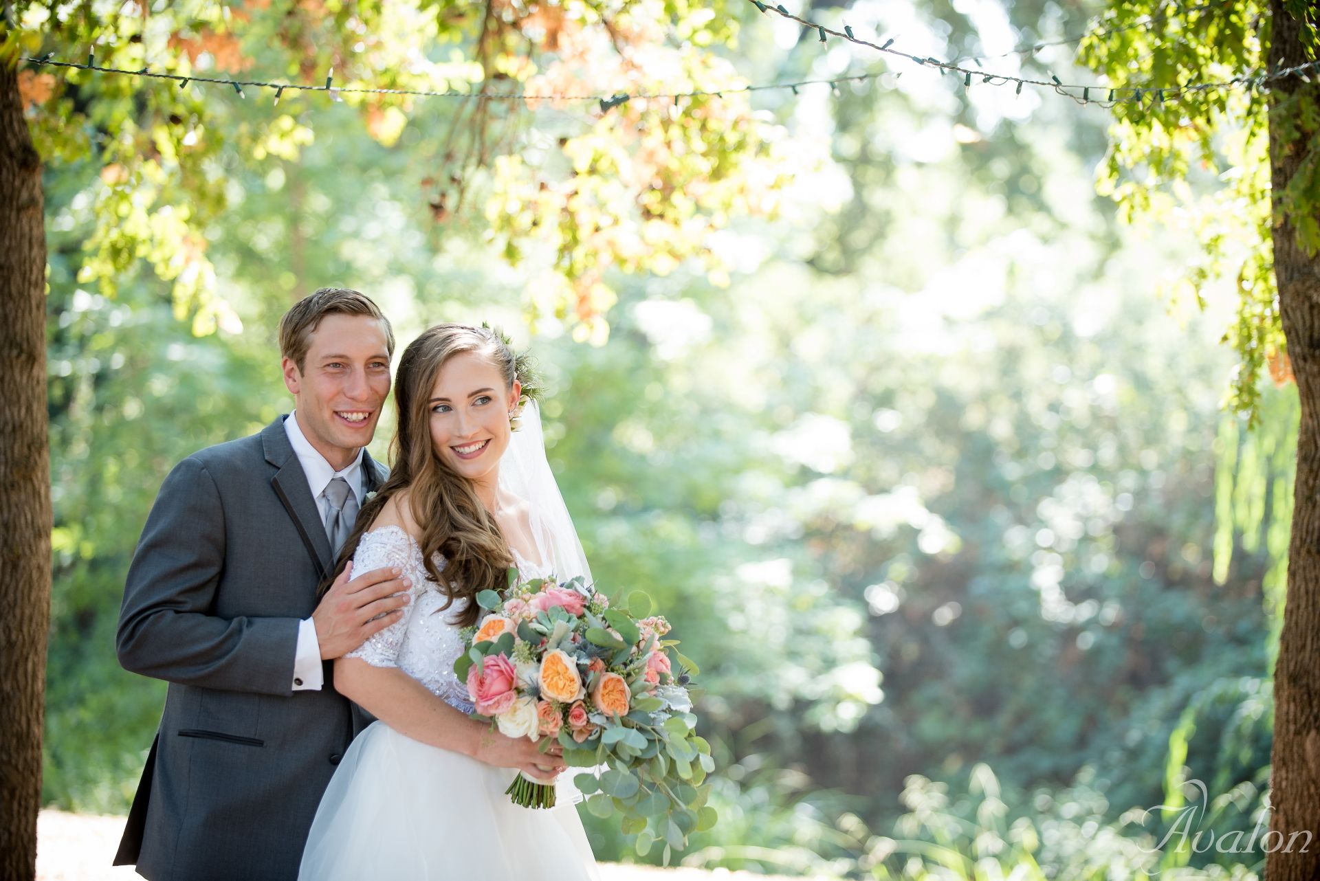 Wedding couple embraces in sunlit garden setting with colorful bouquet.