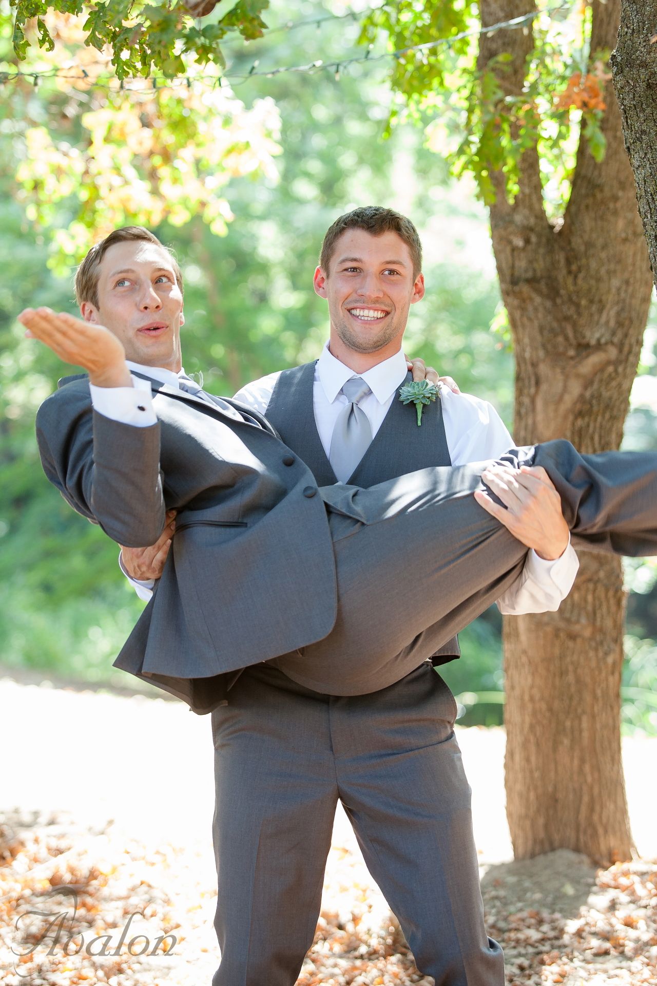 Groomsmen share a playful moment as one carries the other in a bridal-style pose against a sunny outdoor backdrop.