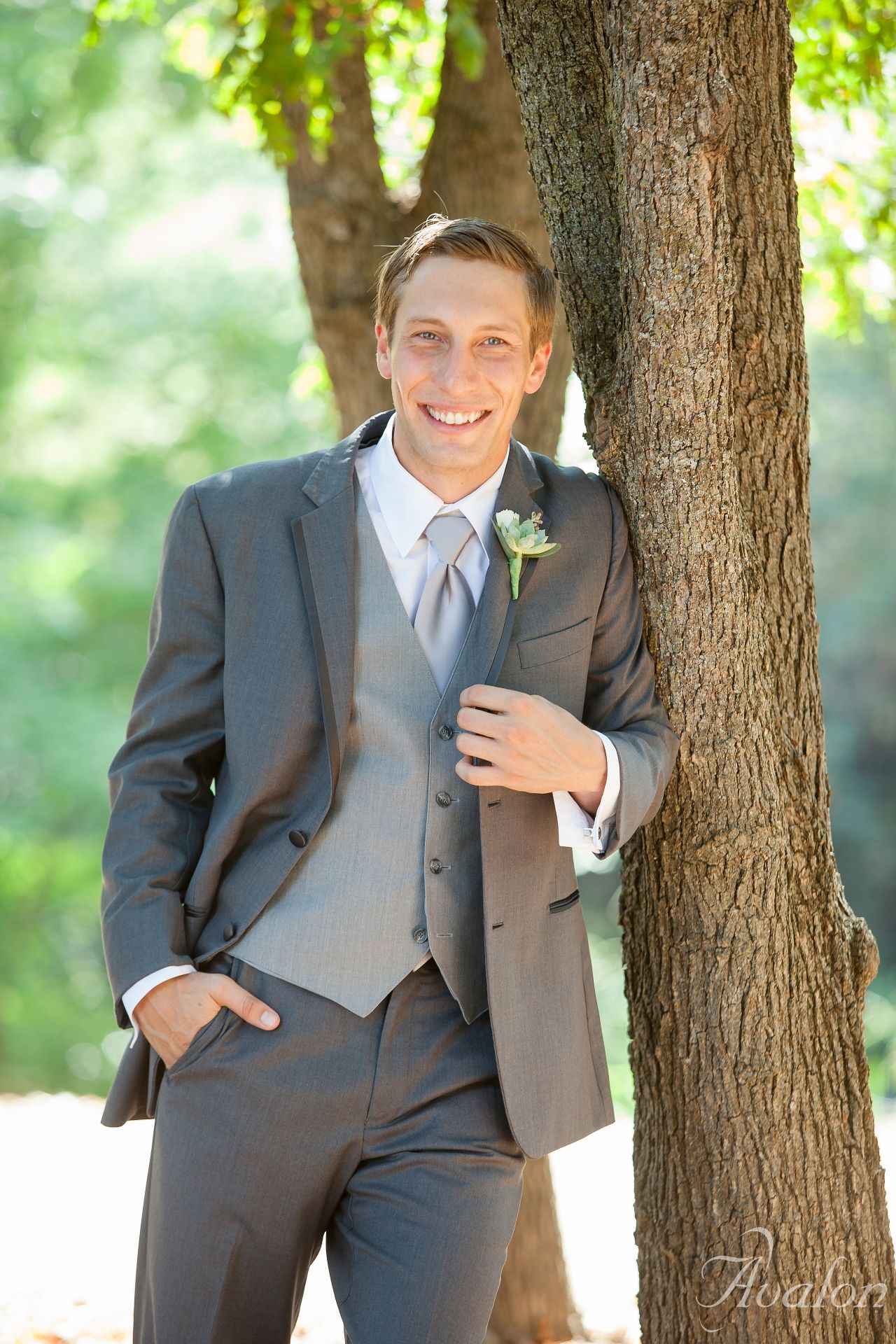 Dapper groomsman in a grey three-piece suit poses confidently against a tree in natural outdoor lighting.