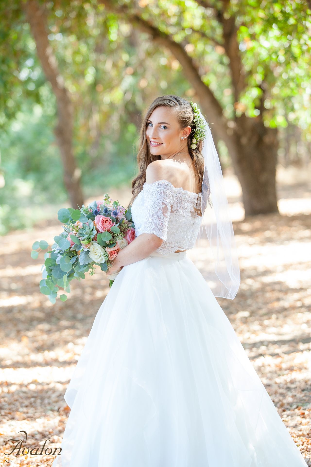 Bride in off-shoulder lace gown poses with elegant bouquet in a sun-dappled woodland setting.