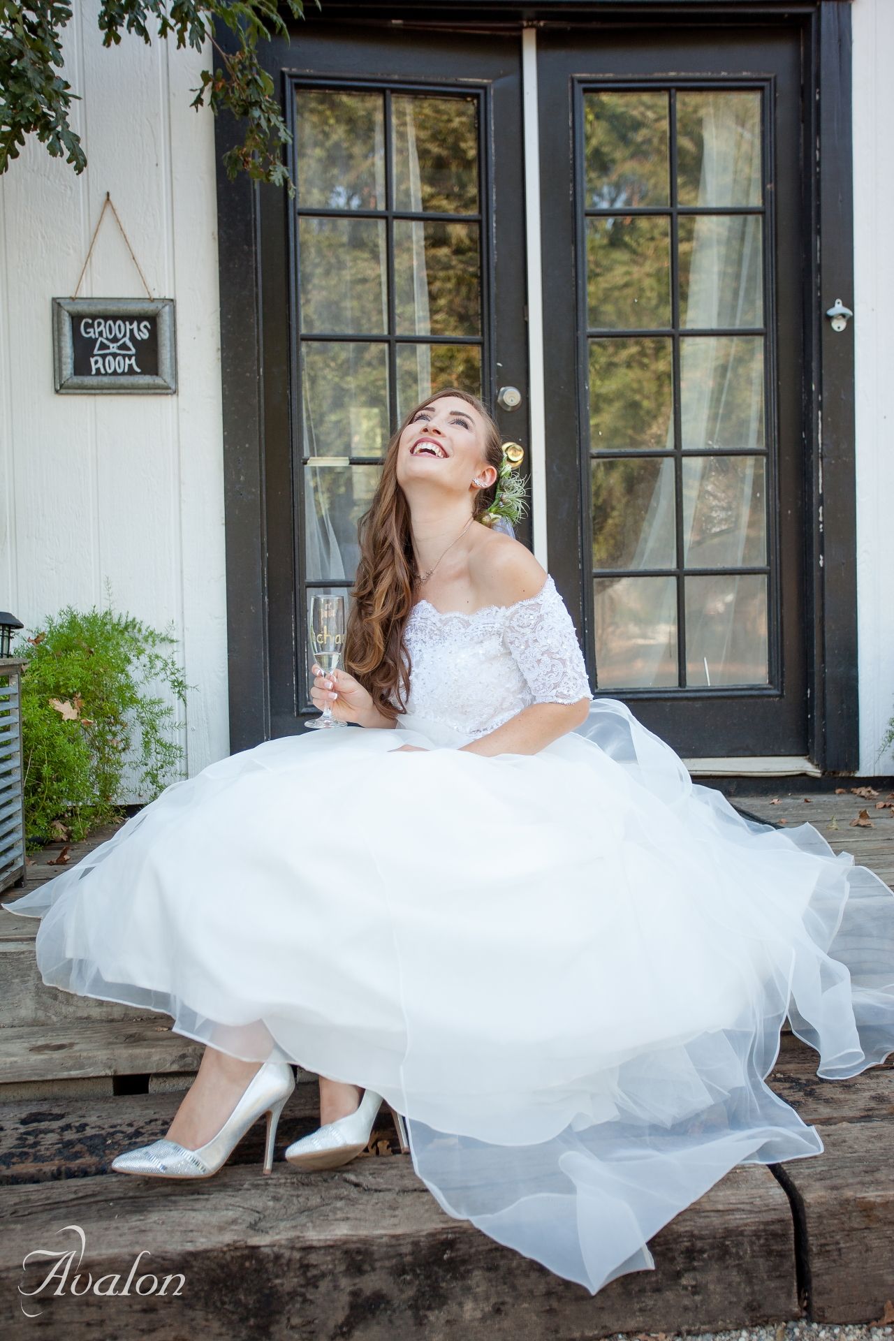 Joyful bride in off-shoulder dress sits laughing on a porch with French doors.