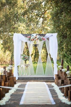 White draped wedding arch decorated with florals leads down an aisle lined with wooden benches.