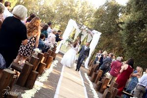 Outdoor wedding ceremony with wooden benches and guests lined up along a sunlit path under trees.