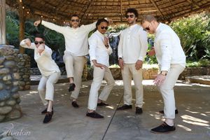 Groomsmen in all-white attire striking fun poses under a thatched roof gazebo.