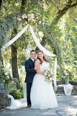 Couple poses under elegant white draped wedding arch decorated with floral arrangements.
