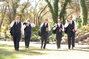 Groomsmen in black vests and ties walk in formation across a sunny outdoor lawn.