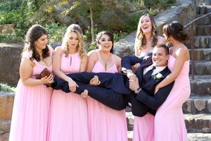 A playful wedding party poses on stone steps with bridesmaids in pink dresses holding a groomsman.