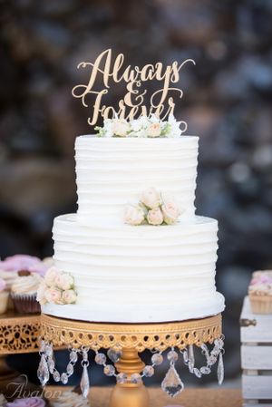 Elegant white wedding cake with 'Always & Forever' topper and blush roses on a gold chandelier stand.