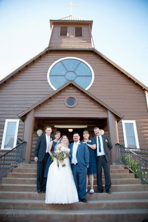 Wedding group photo on church steps with large circular window and cross above the entrance.