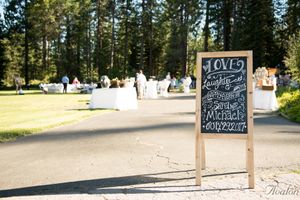 Rustic chalkboard wedding sign stands in grassy outdoor venue with guests in background.