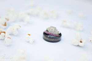 Wedding rings photographed on white surface with diamond engagement ring and black band.