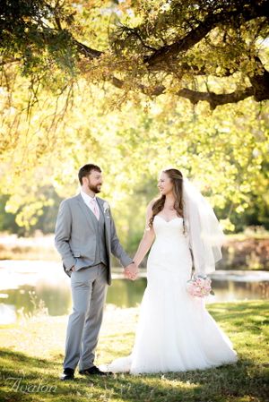 A newlywed couple holds hands under a sunlit tree canopy beside a lake.