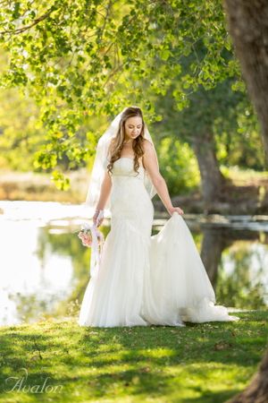 A bride in a flowing white gown poses gracefully by a lake under spring foliage.