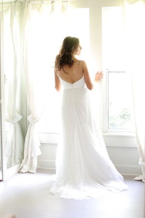 Elegant bridal gown silhouetted against bright window with flowing curtains.
