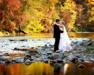 Newlyweds standing on rocky riverbank surrounded by vibrant fall colors.