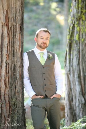 Casual groom portrait in grey vest and lime tie standing between trees.