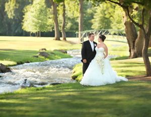 Bride and groom share intimate moment by flowing stream on sunlit grass.