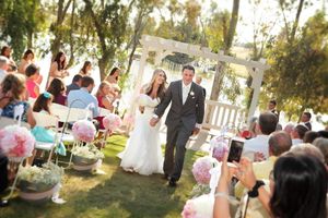 Outdoor garden wedding ceremony with guests taking photos of couple walking down aisle.