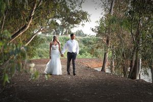 Newlyweds walking hand in hand on rustic dirt path near waterfront.