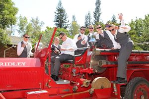 Groomsmen strike playful poses on a vintage red Gridley fire truck at a wedding.