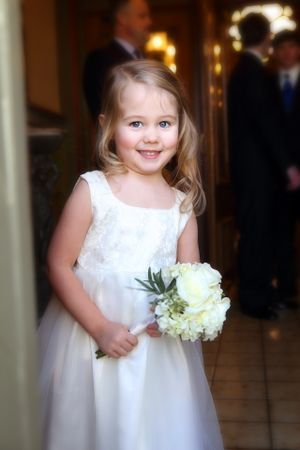 A flower girl in a white dress holds a small bouquet of white flowers in a hallway.