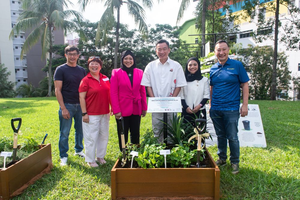 Group of six people posing outdoors near raised garden beds with plants, shovels, and greenery in background.