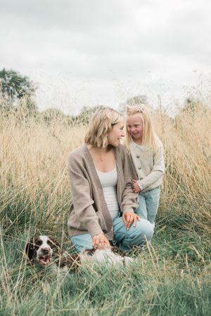 Mother and child share a tender moment in tall grass field with small dog beside them.