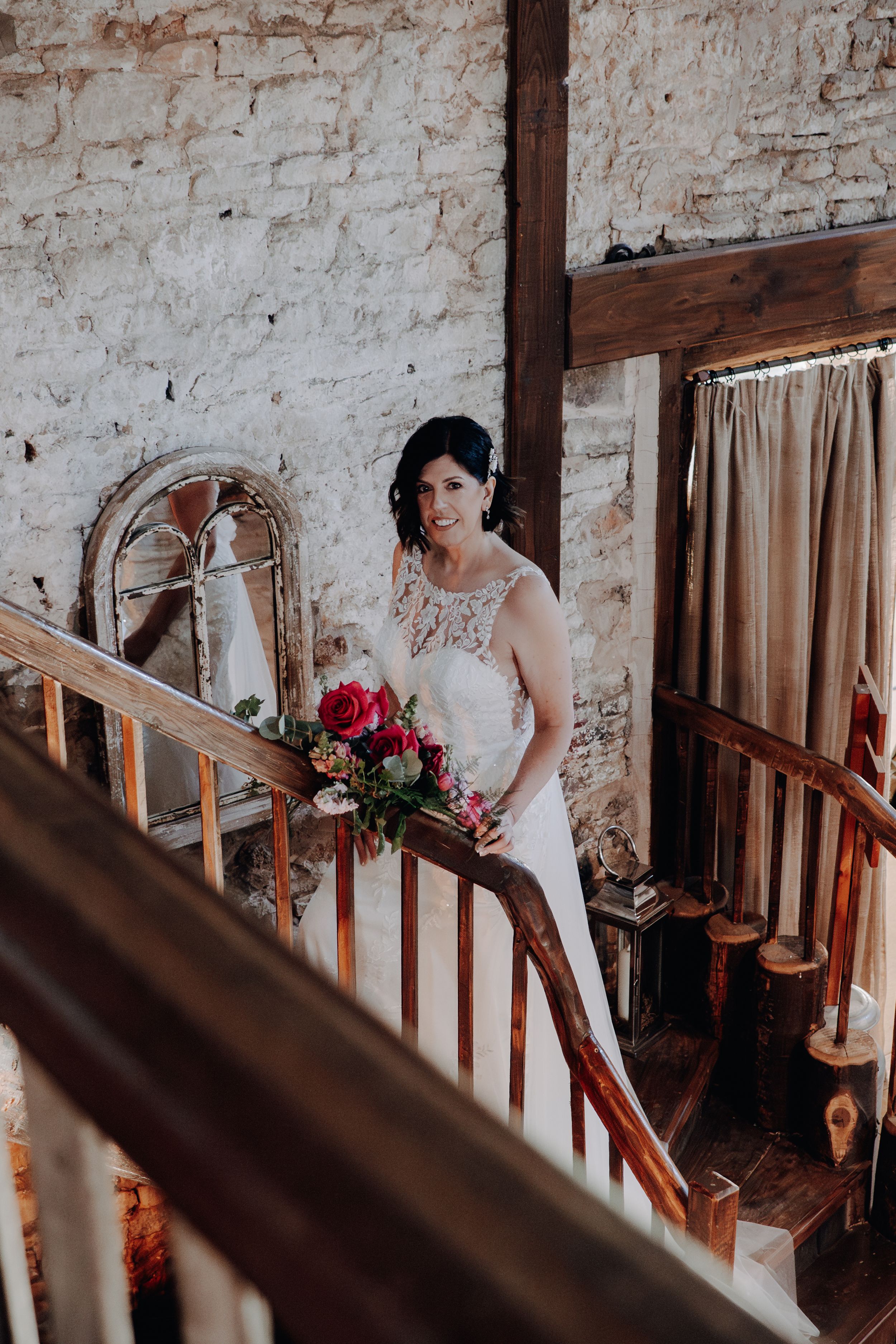 A bride in a lace wedding dress holds a red bouquet while standing on rustic wooden stairs with exposed brick walls.