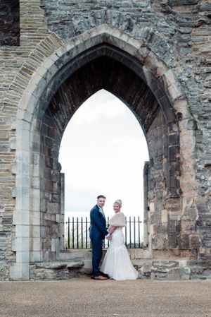 A couple stands framed within a gothic stone archway at a historic castle or cathedral on their wedding day.