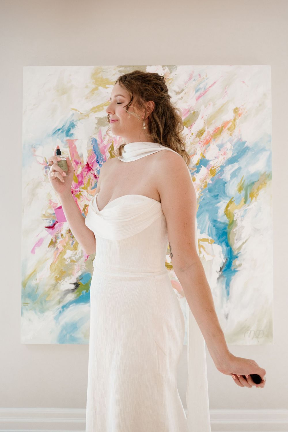 Bride in white halter gown smiling and holding floral bouquet in front of colorful abstract painting.