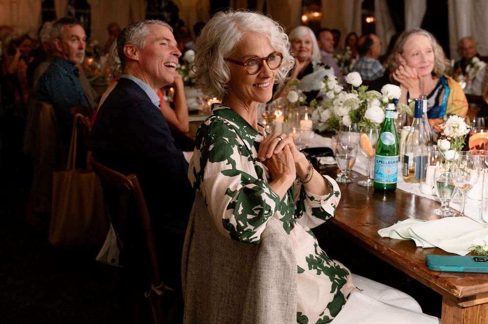 Elderly woman in floral dress clapping joyfully at an elegant dinner party with guests seated at candlelit tables.